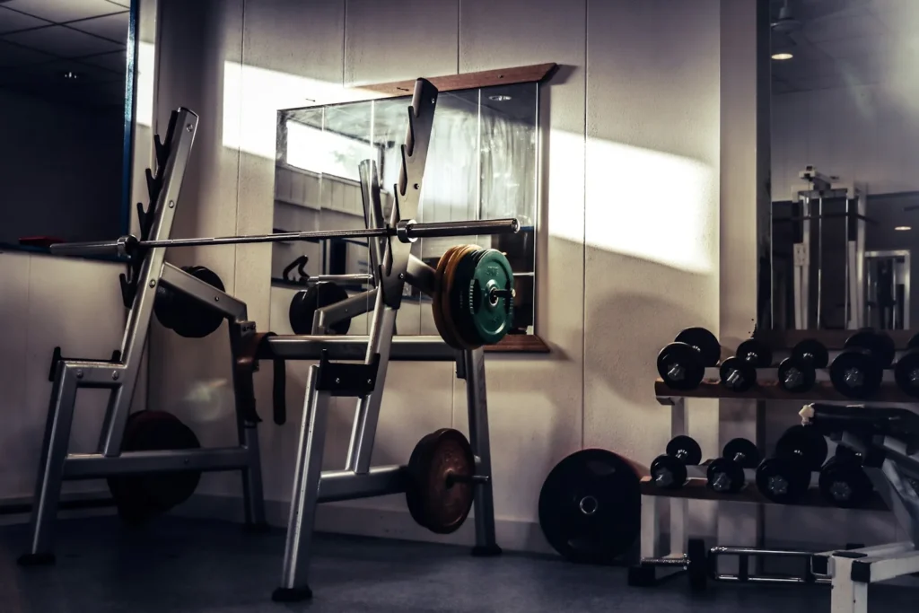 Salle de musculation avec un rack à squat, une barre chargée et des haltères, traversée par un rayon de soleil oblique sur le mur